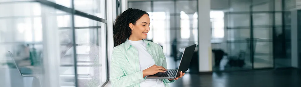 Uma jovem mulher sorridente, com cabelos cacheados escuros, está em pé em um ambiente de escritório moderno e iluminado, utilizando um laptop preto. Ela veste uma camiseta branca e uma camisa verde clara listrada aberta. Ao seu redor, paredes de vidro refletem a luz e mostram partes de outros espaços de trabalho. A cena sugere um ambiente profissional dinâmico, onde a jovem pode estar trabalhando em um contrato de estágio ou outras tarefas relacionadas aos seus estudos ou início de carreira.