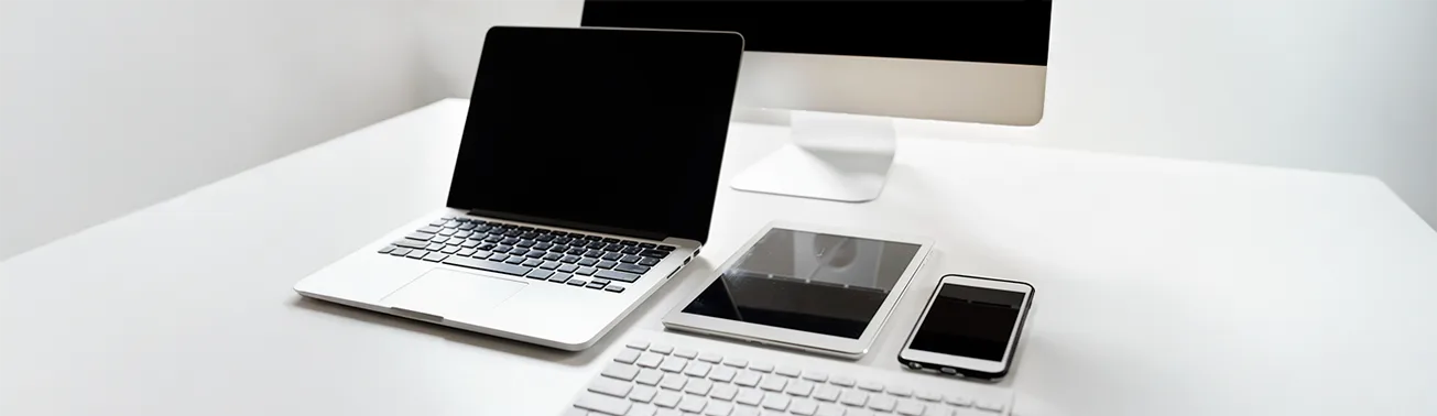 Uma estação de trabalho minimalista com um laptop, um tablet e um smartphone sobre uma mesa branca. A composição de dispositivos eletrônicos sugere ferramentas de produtividade e conectividade, destacando a utilidade de um aplicativo para escanear documentos para digitalização e organização de informações no ambiente de trabalho moderno. Um teclado branco e um monitor também estão presentes.