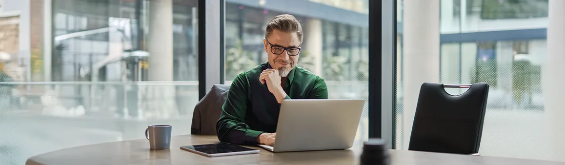 Um homem de óculos, vestindo uma camisa verde, está sentado em um escritório moderno com grandes janelas, olhando pensativamente para seu laptop. Ele tem a mão no queixo, o que pode indicar que está revisando documentos importantes ou considerando decisões complexas, como um distrato de contrato. Um tablet e uma caneca estão na mesa ao seu lado.