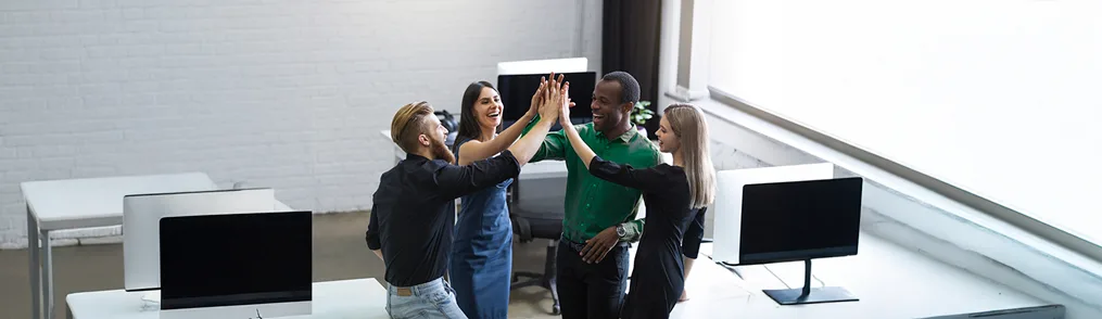 Quatro colegas de trabalho sorridentes, dois homens e duas mulheres, estão em um escritório moderno, com as mãos unidas em um "high-five" no centro, celebrando o sucesso. A cena representa um trabalho em equipe eficaz e a conquista de metas, que pode ser aprimorada por metodologias de organização e produtividade, como o conceito de "O que é 5S", promovendo um ambiente de trabalho otimizado. O local possui mesas brancas e monitores pretos, com grandes janelas à direita.