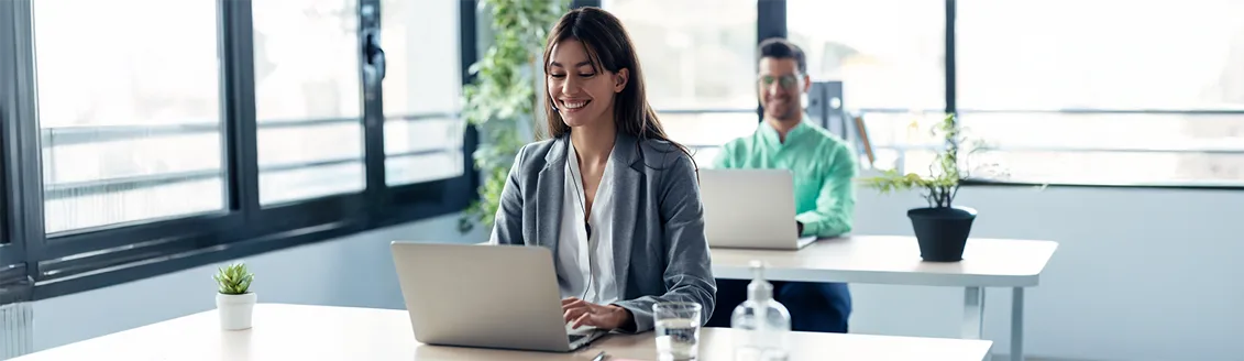 Uma mulher sorridente em um blazer cinza trabalha em seu laptop em um escritório bem iluminado, com plantas e grandes janelas ao fundo. Um colega de trabalho também está visível ao fundo, usando um laptop. A imagem transmite um ambiente de trabalho profissional e organizado, onde o uso de documentos formais, como um papel timbrado, seria comum para comunicação e padronização.
