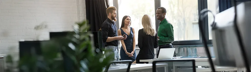 Quatro colegas de trabalho estão engajados em uma conversa animada em um escritório moderno, com grandes janelas ao fundo. Eles parecem estar colaborando e trocando ideias, possivelmente discutindo estratégias para alcançar quick wins e obter resultados rápidos. A atmosfera é de cooperação e proatividade.