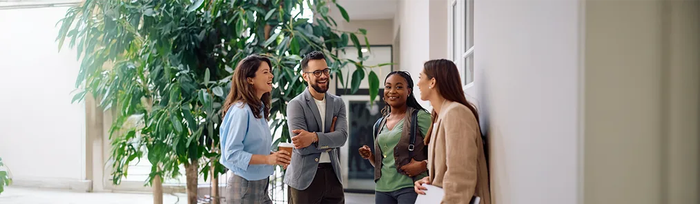 Quatro colegas de trabalho sorridentes, dois homens e duas mulheres, estão conversando em um corredor iluminado por luz natural e rodeado por plantas grandes. Eles estão engajados em uma discussão animada, demonstrando bom relacionamento e a prática da escuta ativa mútua enquanto interagem.