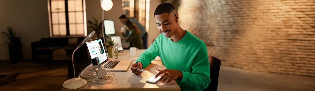 Um homem sorridente, vestindo uma blusa verde, está sentado em uma mesa de escritório, usando um smartphone e segurando um cartão de crédito. A cena sugere que ele está gerenciando uma conta ou realizando uma compra, possivelmente relacionada a serviços de telecom, com um colega de trabalho ao fundo.