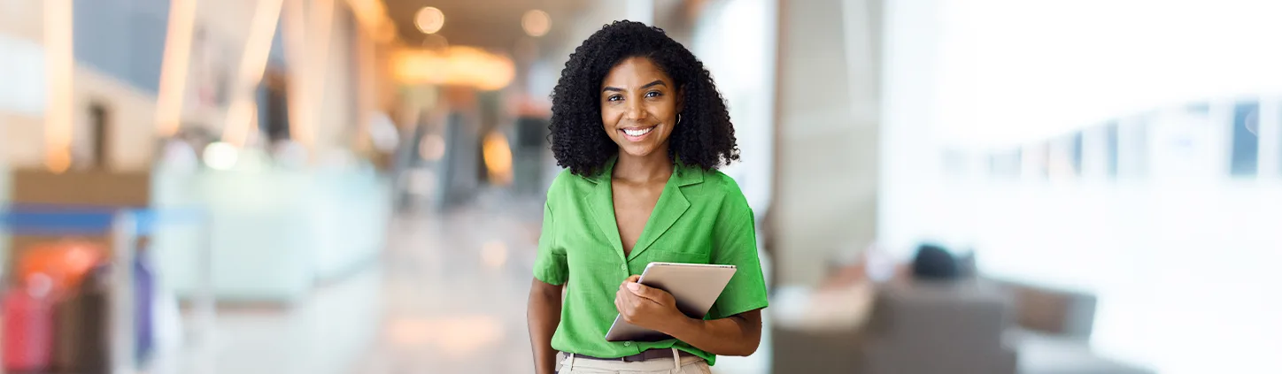 Uma mulher sorridente, com cabelos cacheados e uma blusa verde, segura um tablet em um ambiente moderno e bem iluminado. A imagem a retrata como uma profissional confiante e pode simbolizar a facilidade de assinar um contrato de marketing digital de forma eletrônica.