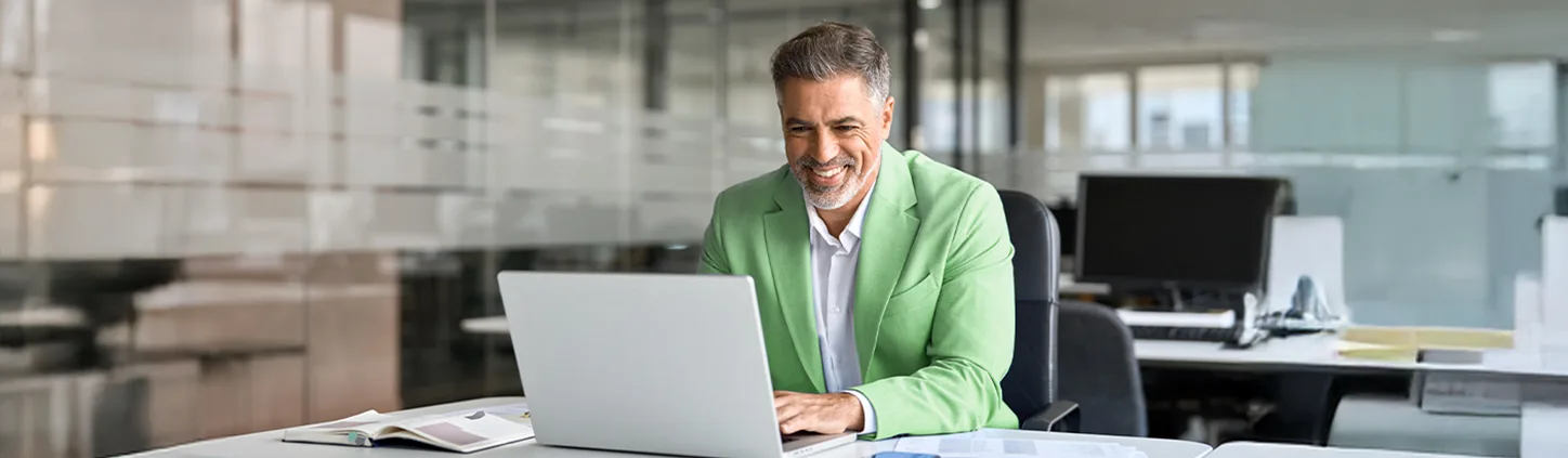 Um homem sorridente de meia-idade, com um blazer verde, está sentado em sua mesa em um escritório moderno, usando um laptop. A imagem representa a crescente integração da tecnologia no direito, com o profissional utilizando ferramentas digitais para otimizar seu trabalho.
