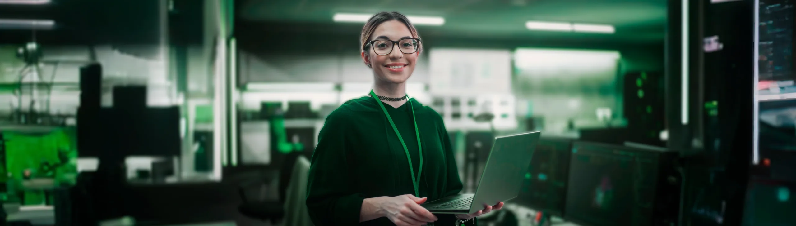 Uma mulher sorridente com óculos e suéter verde está segurando um laptop em um ambiente de tecnologia, com múltiplos monitores ao fundo. A cena representa a acessibilidade e o empoderamento proporcionados por plataformas de low-code e no-code, permitindo que profissionais criem soluções digitais.