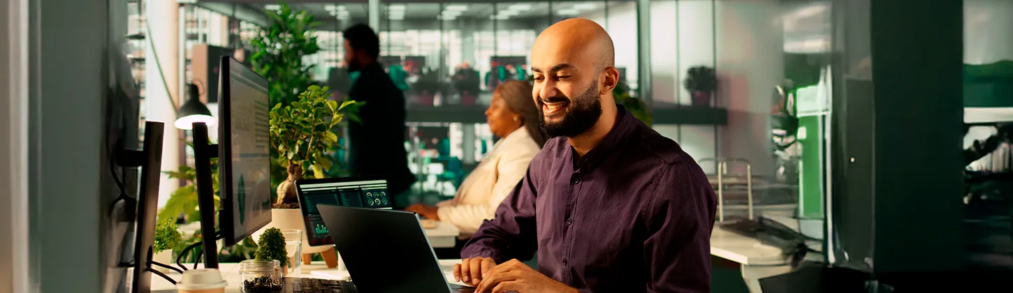 Um profissional de TI sorridente trabalha em seu laptop em um escritório moderno, gerenciando seu contrato para profissionais de TI.