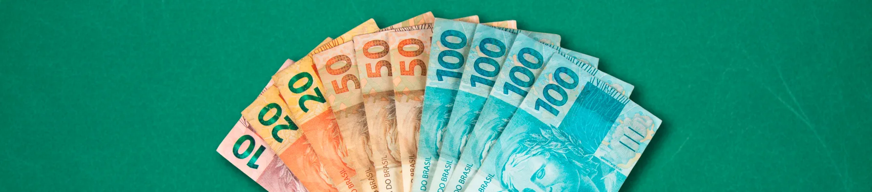 Várias cédulas de Real (10, 20, 50 e 100) abertas em leque sobre fundo verde, ilustrando os impactos econômicos da Reforma Tributária.